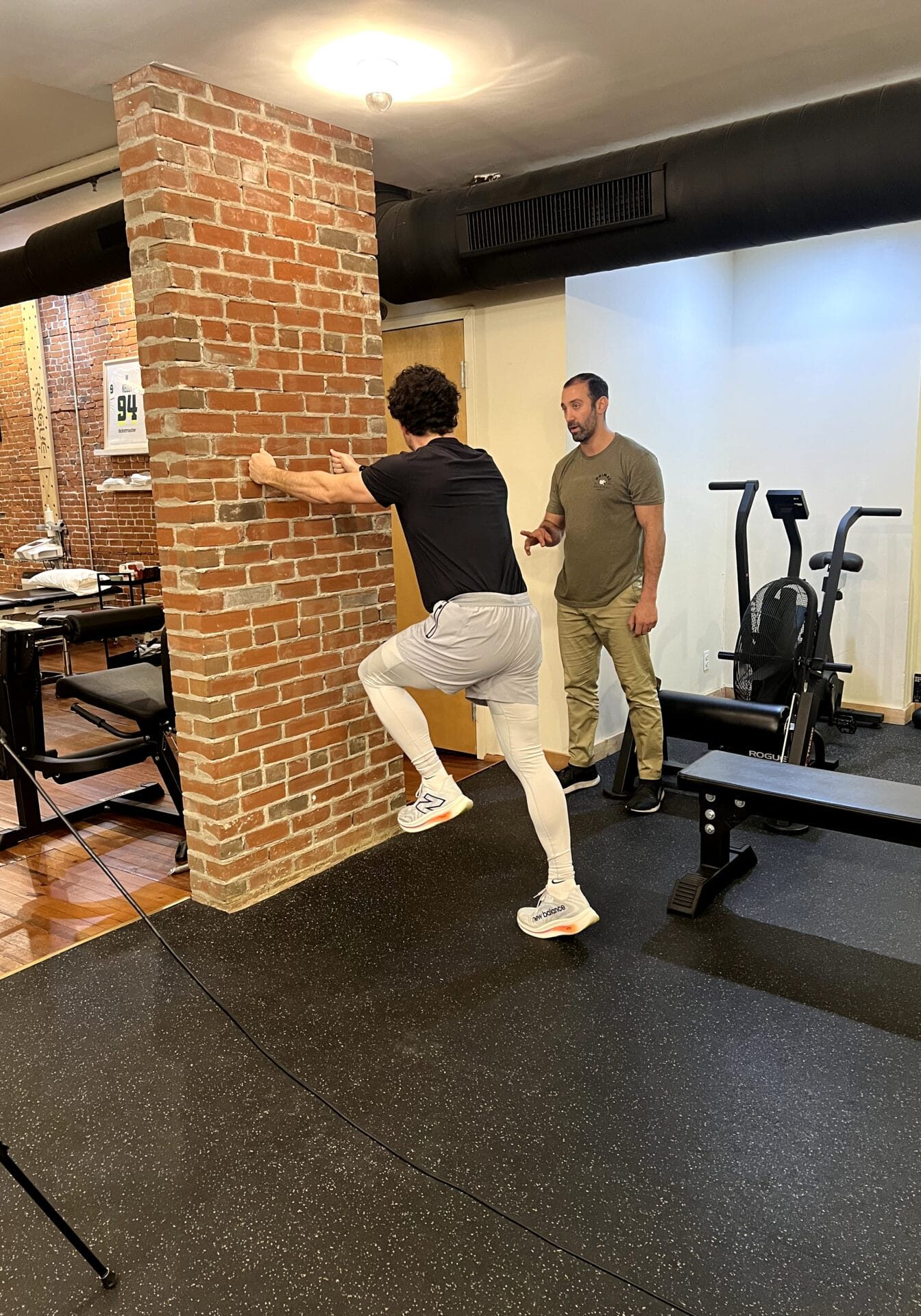 Athlete performs a single-leg wall drill against a brick pillar while a physical therapist observes in a treatment facility, focusing on balance and lower-body strength.