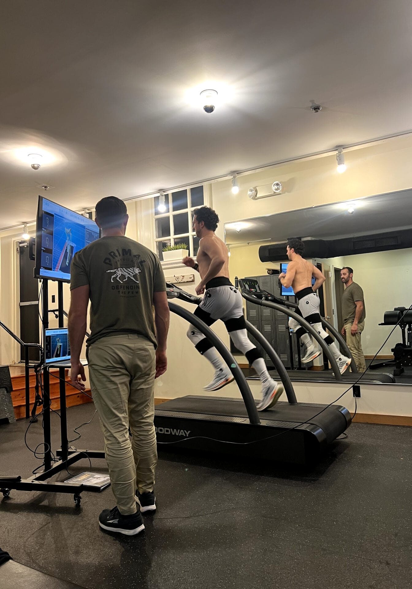 Physical therapist monitors an athlete running on a curved treadmill during a gait analysis session, with performance data displayed on a screen in a treatment facility.