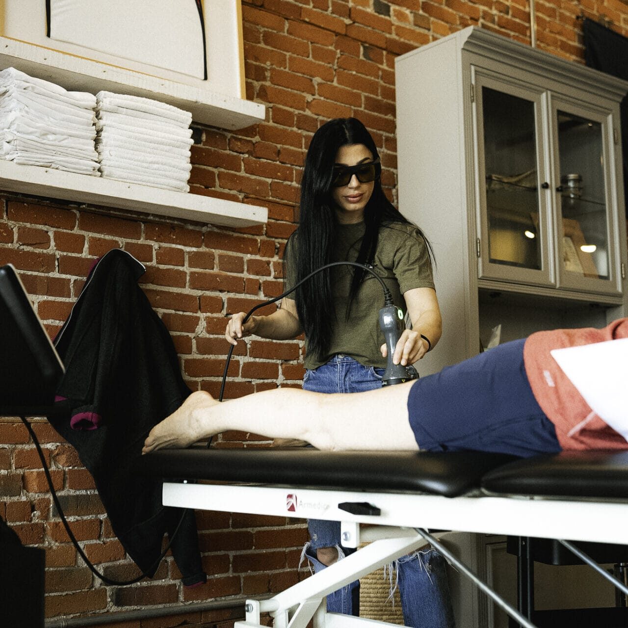 Physical therapist wearing protective glasses performing laser therapy on a patient’s leg in a modern clinic with exposed brick walls.