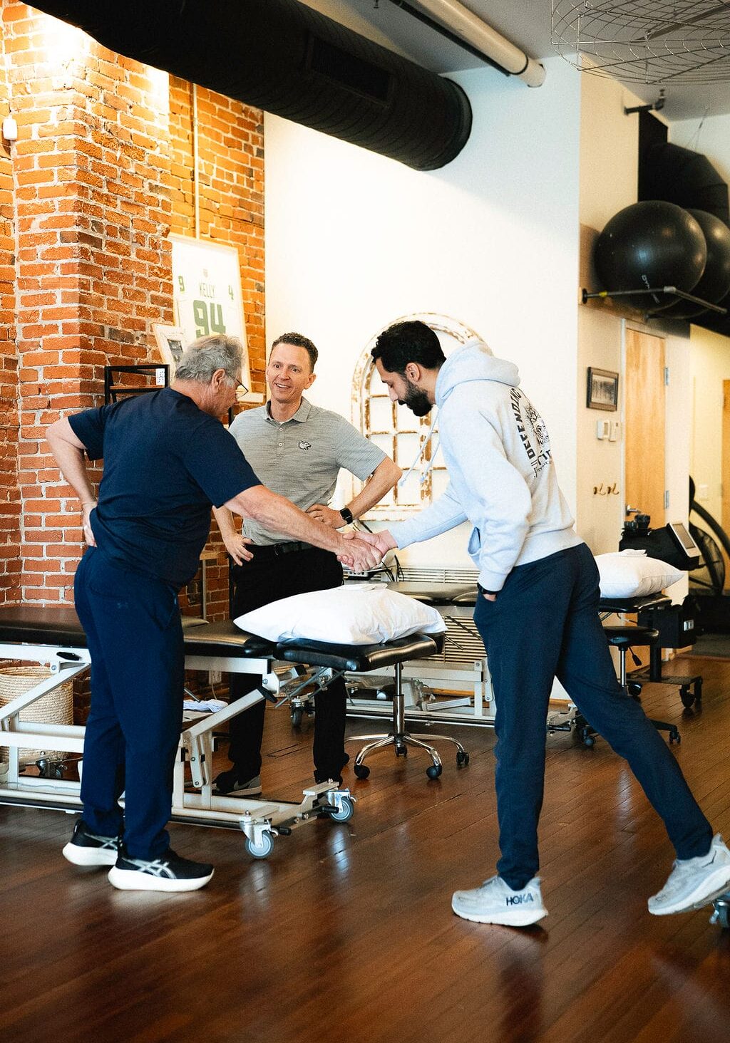 Physical therapy session in a gym-style clinic, with a therapist coaching two clients during a hands-on movement assessment beside treatment tables and pillows.