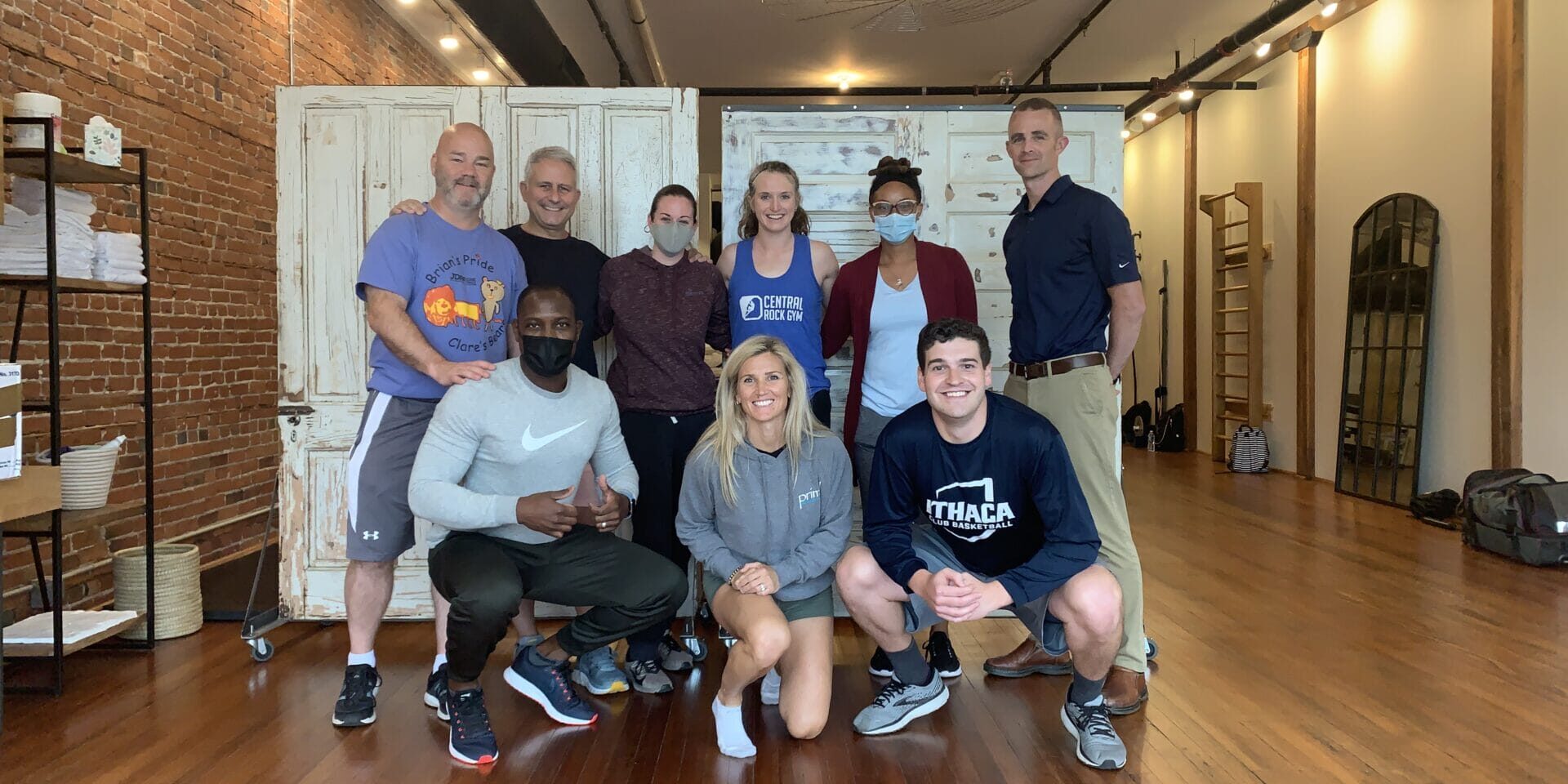 Primal Physical Therapy team group photo in a bright fitness studio with exposed brick, wooden floors, and rustic doors; several team members wear masks.