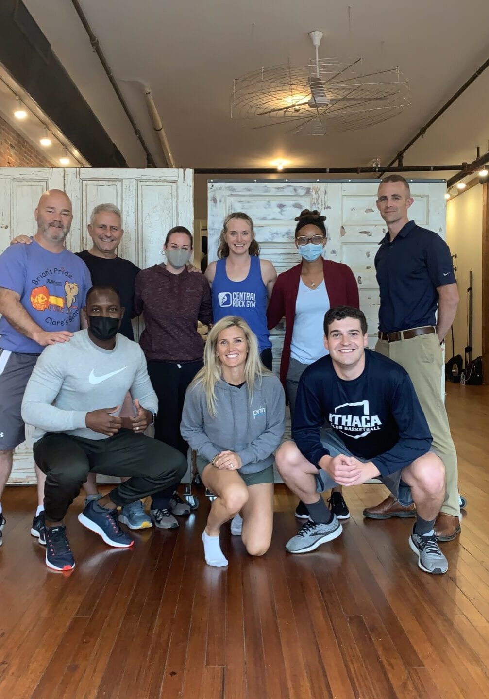 Primal Physical Therapy team group photo in a bright fitness studio with exposed brick, wooden floors, and rustic doors; several team members wear masks.