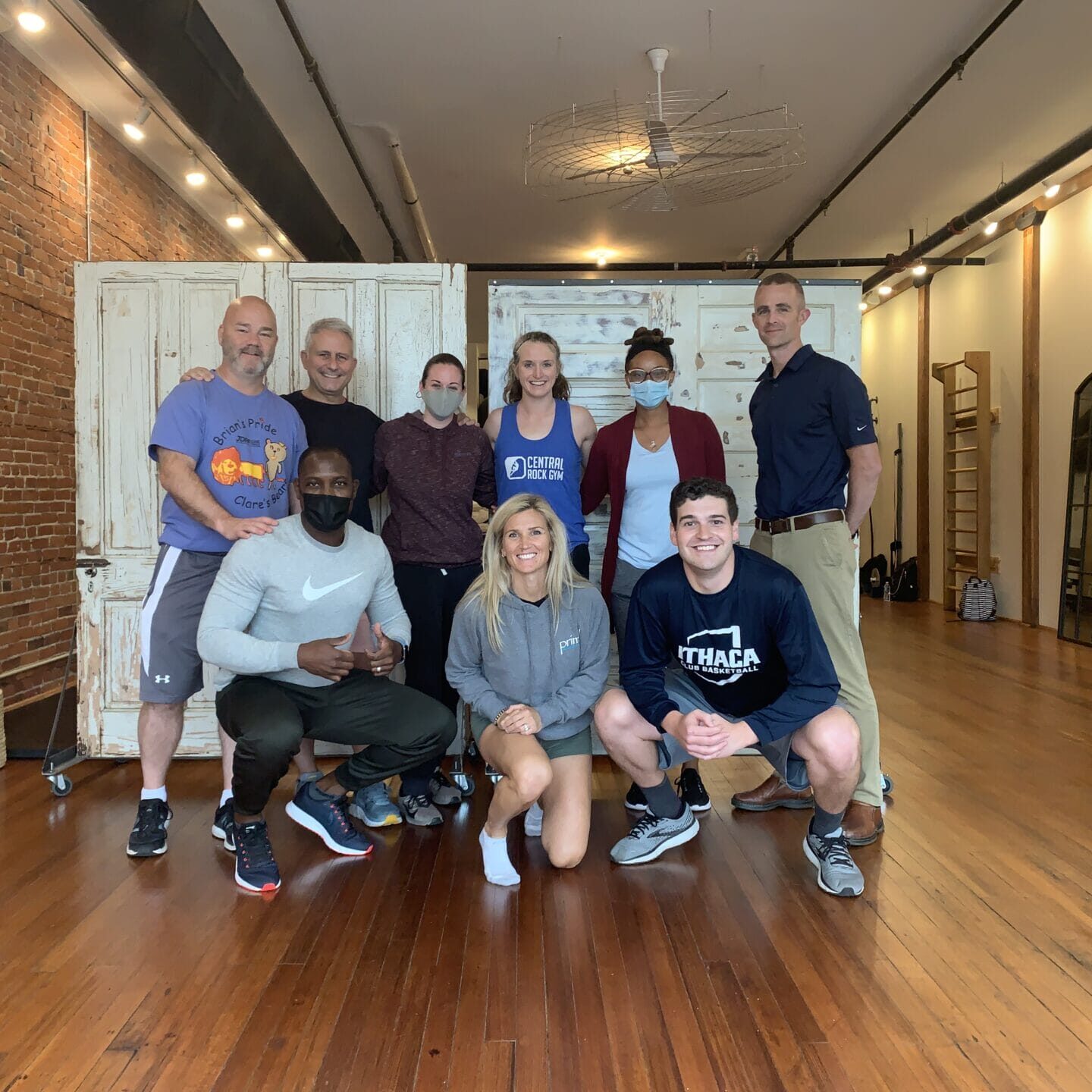 Primal Physical Therapy team group photo in a bright fitness studio with exposed brick, wooden floors, and rustic doors; several team members wear masks.